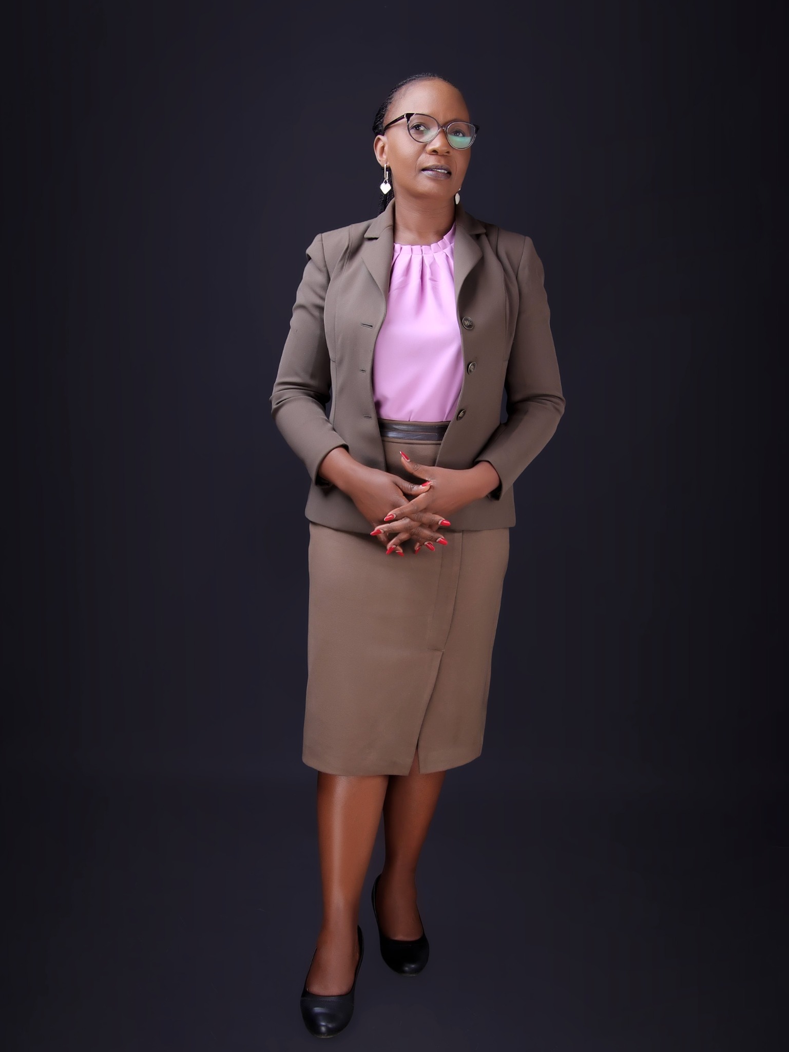 Dr. Victoria Wanambiro in a brown suit and pink blouse, standing against a dark background